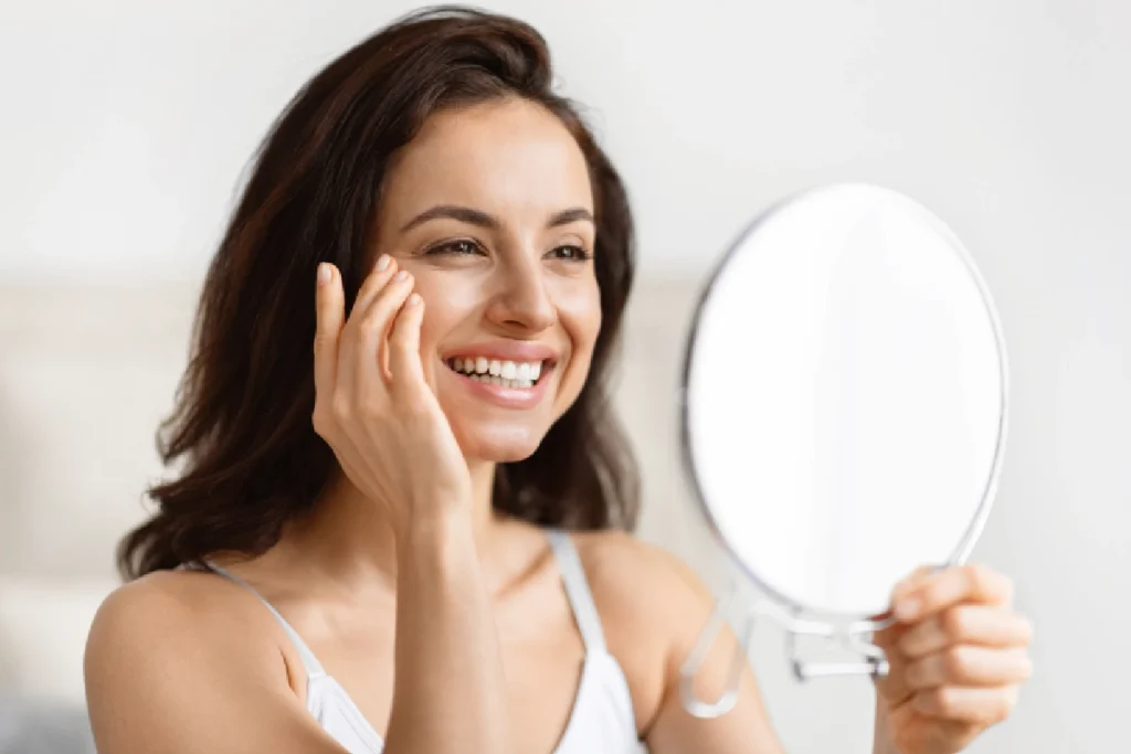 Woman with healthy, glowing skin smiling into a mirror after applying SkinCeuticals C E Ferulic antioxidant serum for environmental aging protection and skin barrier support.