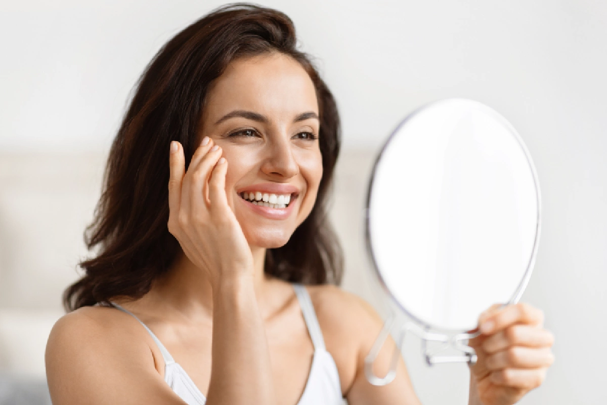 Woman with healthy, glowing skin smiling into a mirror after applying SkinCeuticals C E Ferulic antioxidant serum for environmental aging protection and skin barrier support.